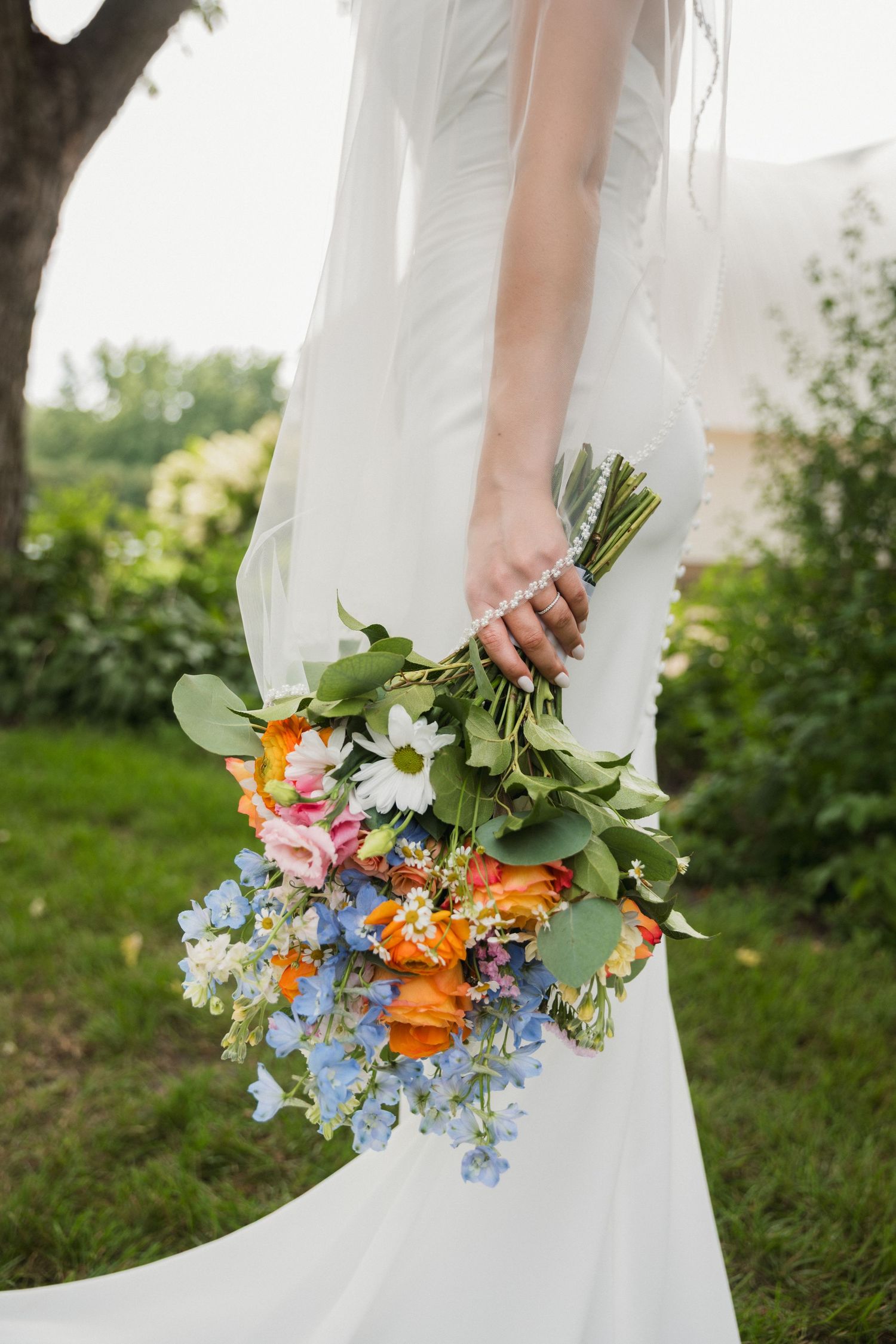 Bride in white gown holding colorful wildflower bouquet with orange, blue, and pink blooms outdoors.