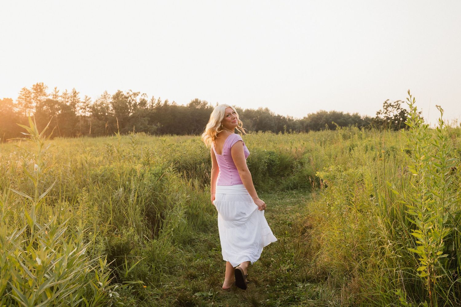 A young blonde woman in a pink top and white skirt walks through a sunlit green meadow at golden hour.