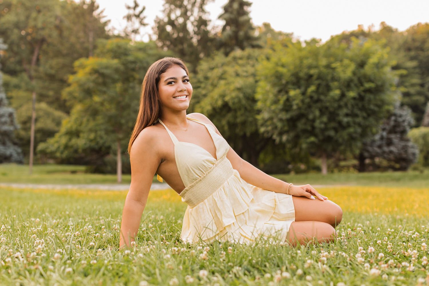 Young woman in a cream dress sits in a meadow of white wildflowers surrounded by lush green trees in summer.