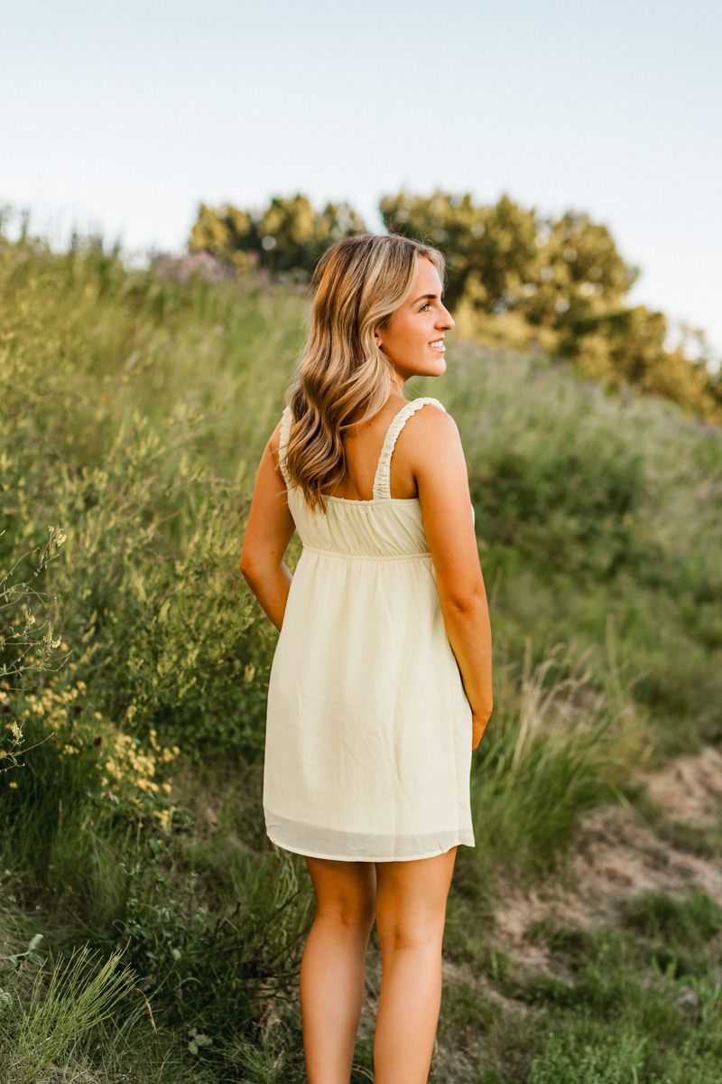 Woman in cream sundress standing in golden meadow at sunset with trees in background.