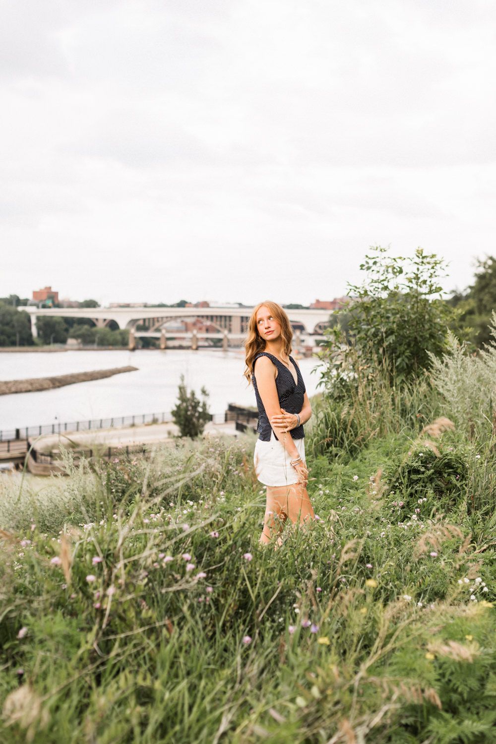 Young woman in navy top and white shorts poses on grassy riverbank with bridge and waterfront in background.
