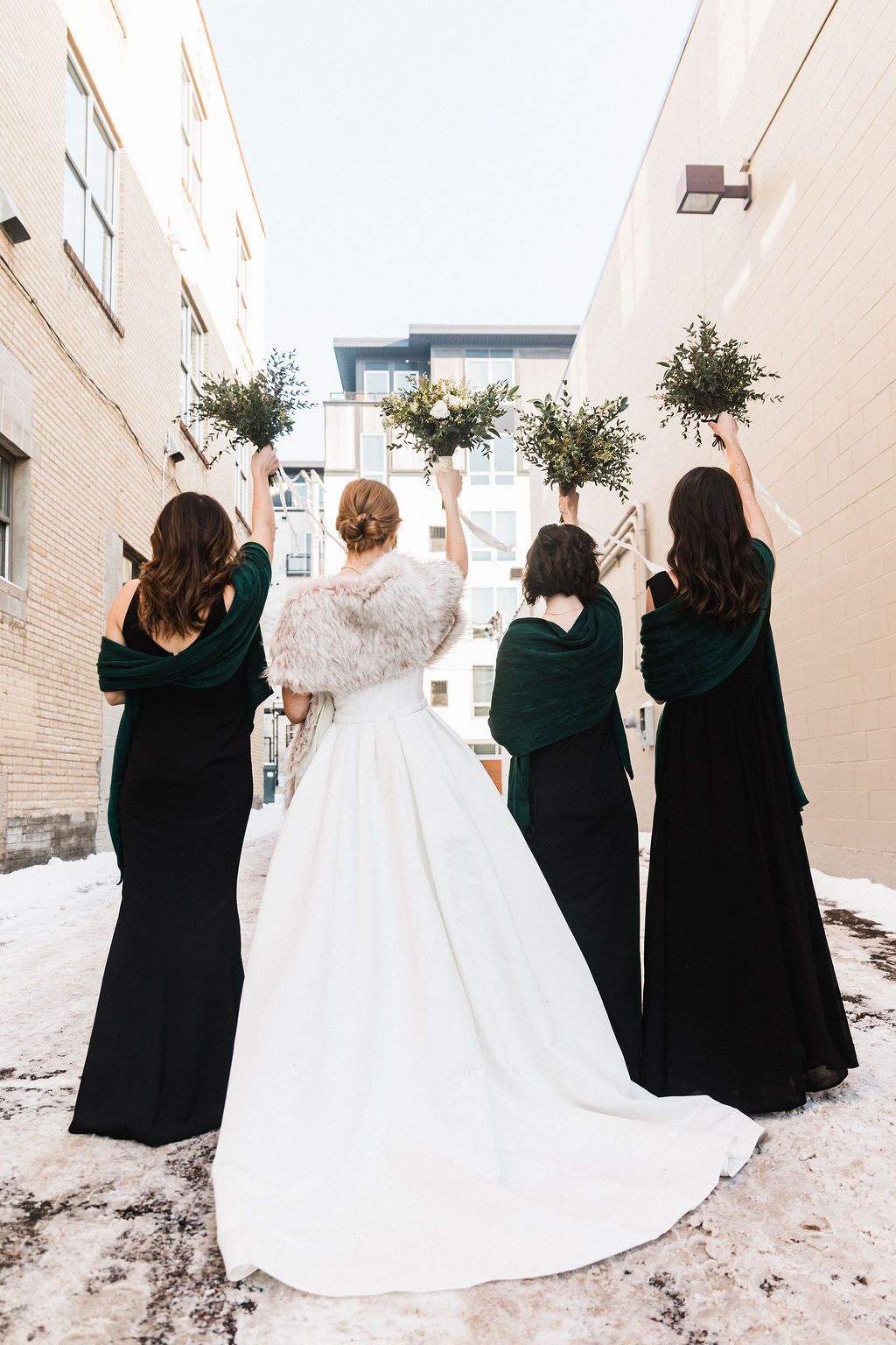 Bridesmaids in black dresses pose with bride in white gown against snowy outdoor backdrop.