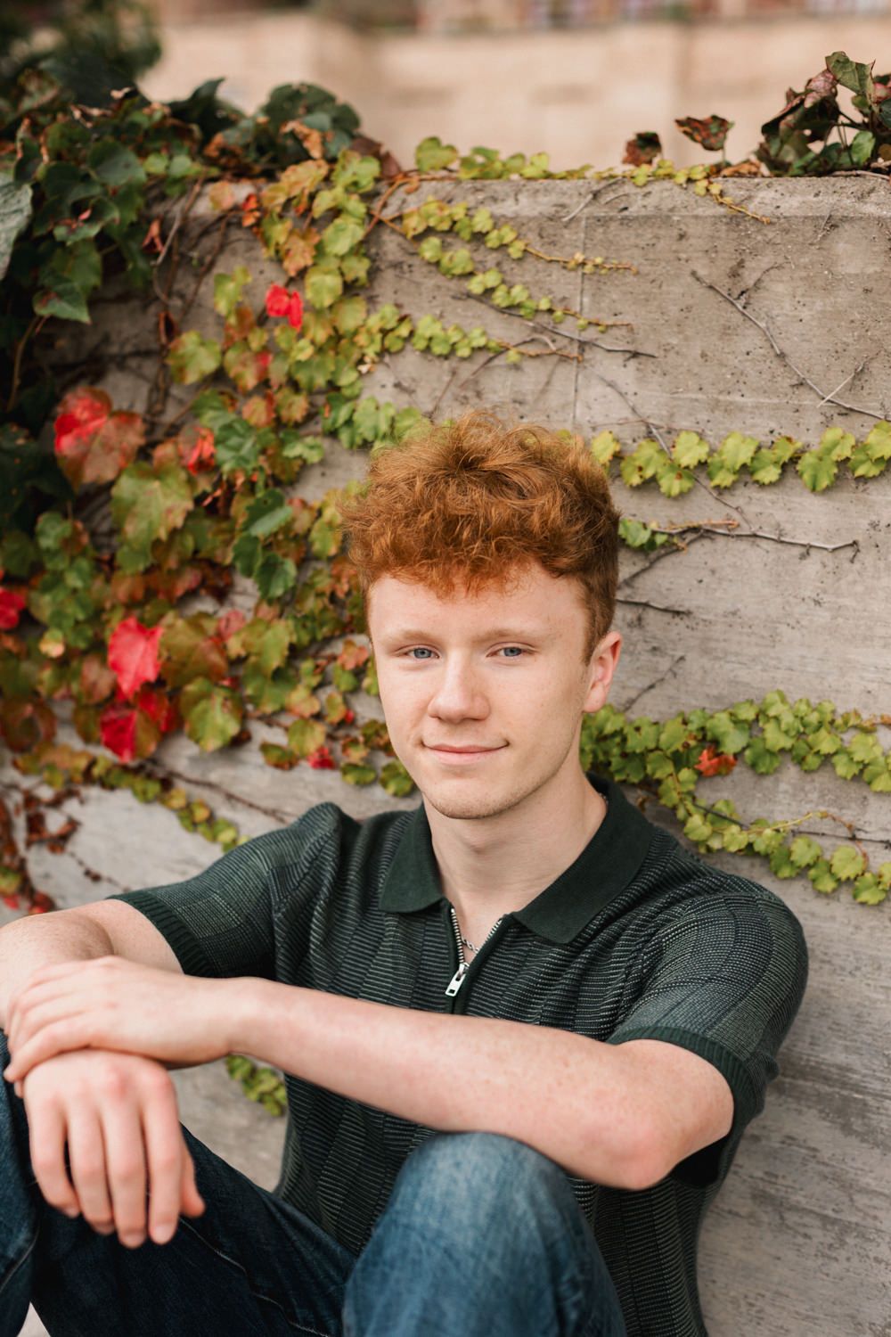 Portrait by stone wall with green climbing vines and red flowers in the background.