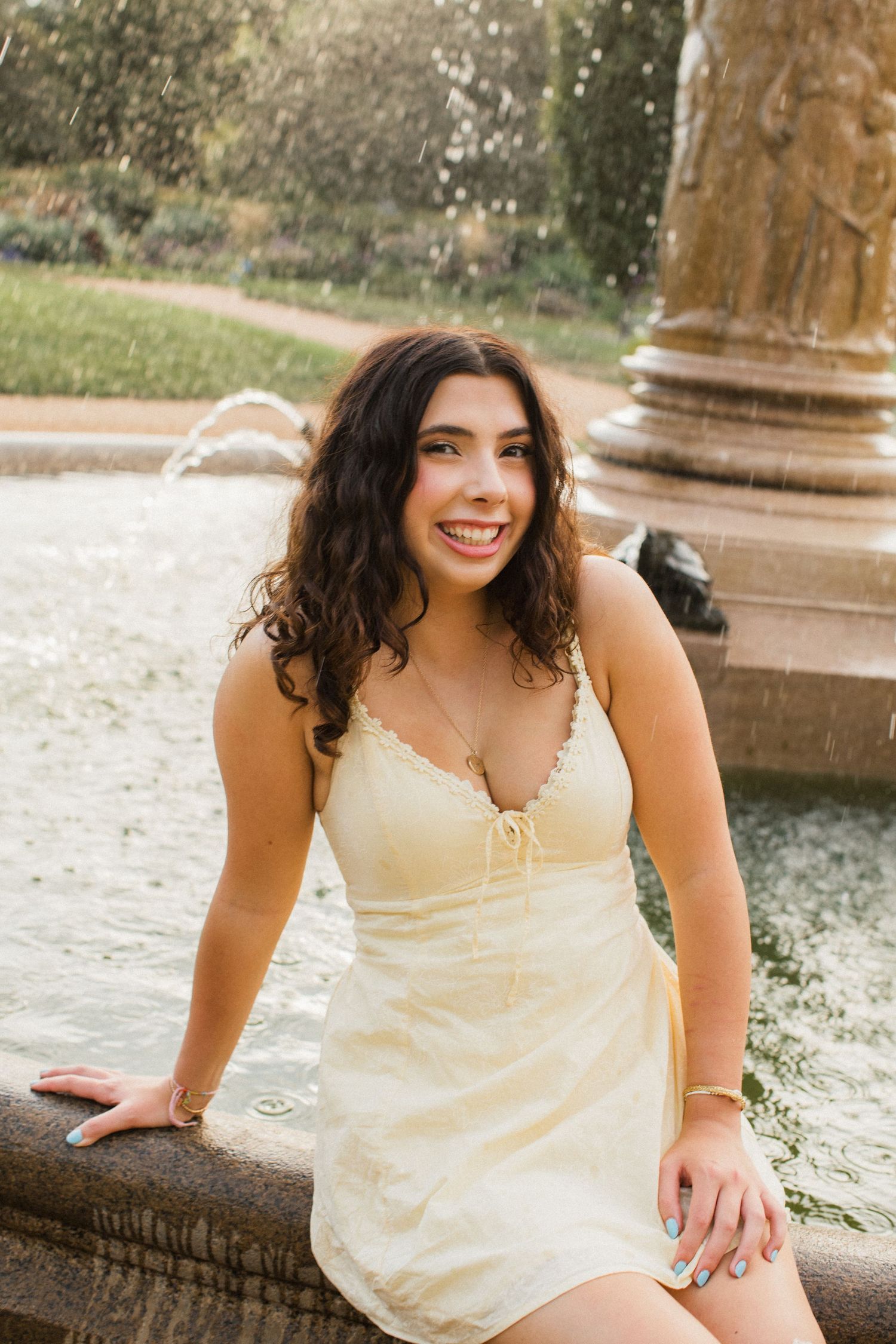 Young woman in a cream sundress smiling by a stone fountain in a sunny park setting.
