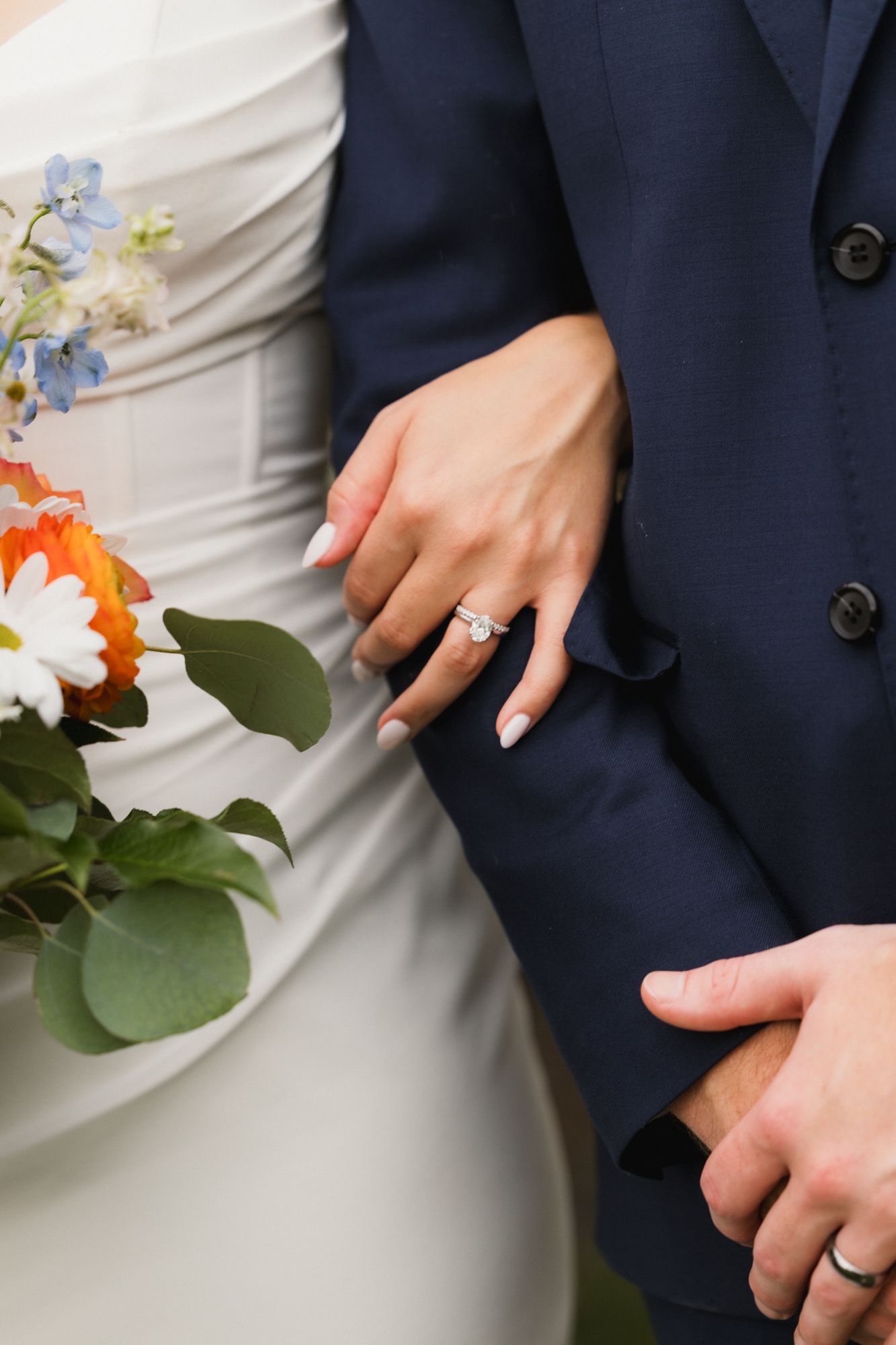 Bride and groom standing together, showcasing diamond engagement ring and wedding band with a colorful bridal bouquet.