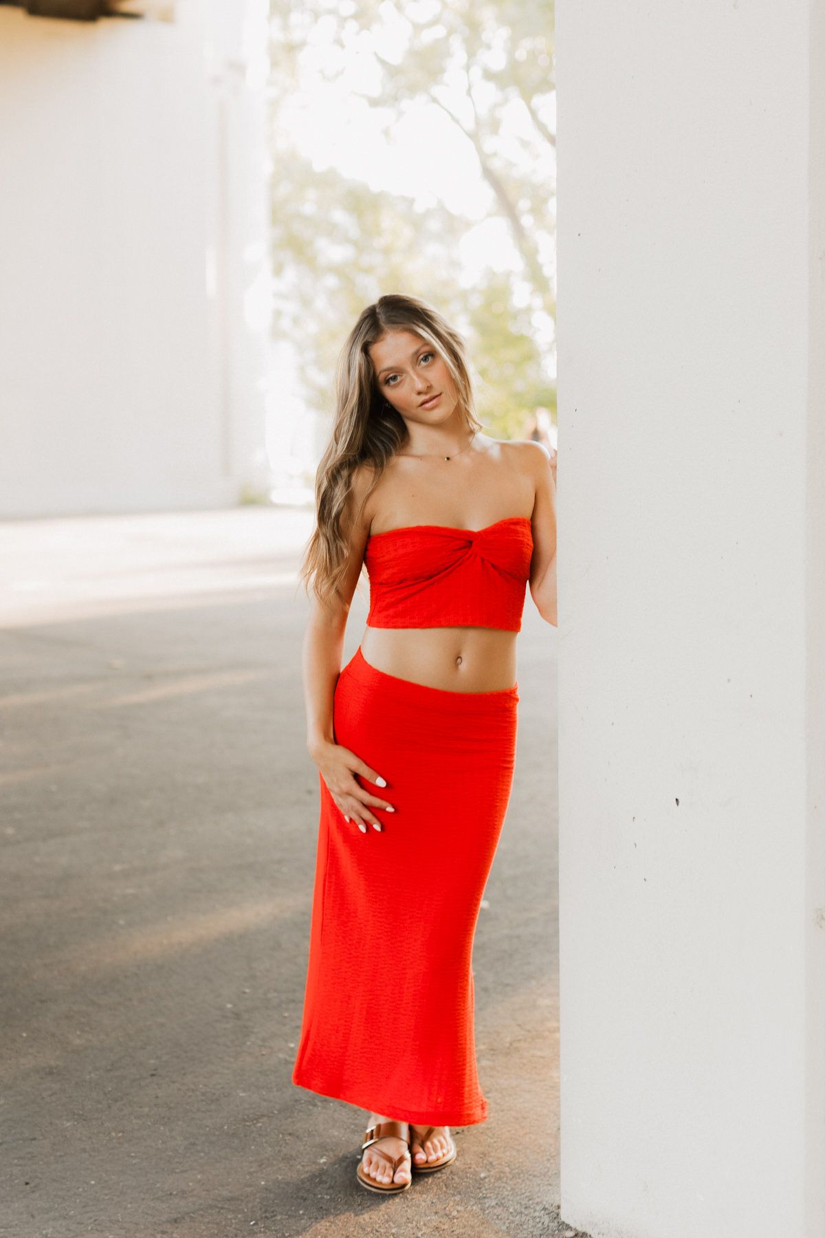 Woman in red strapless crop top and maxi skirt posing outdoors by white column.