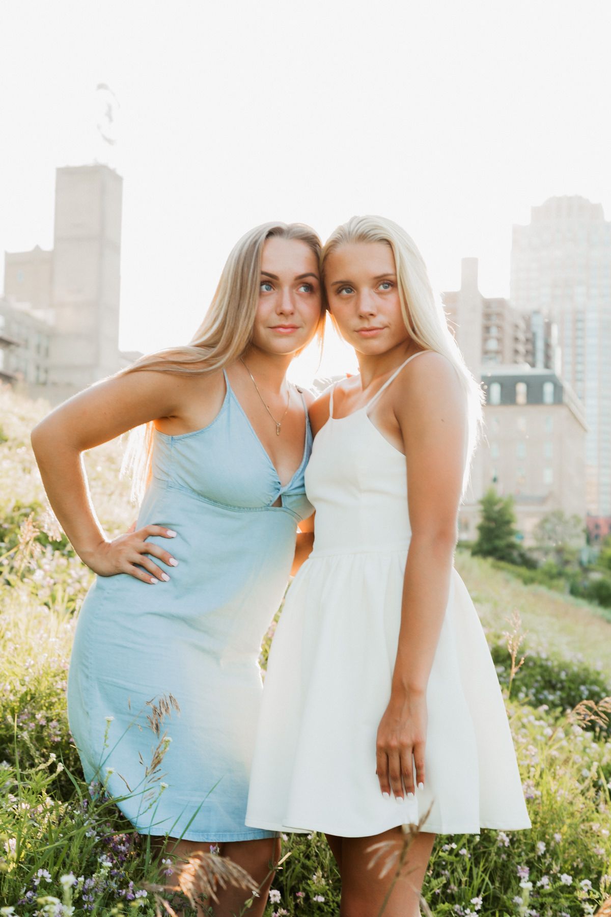 Two blonde women in light summer dresses pose together in a sunlit urban garden.