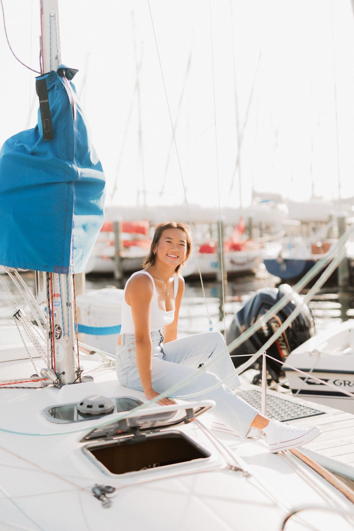 Happy woman in white outfit sitting on a sailboat deck at a sunny marina.
