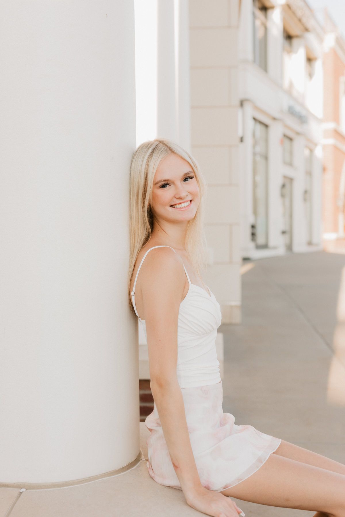 Smiling blonde teen girl in white dress leaning against a white column outdoors.