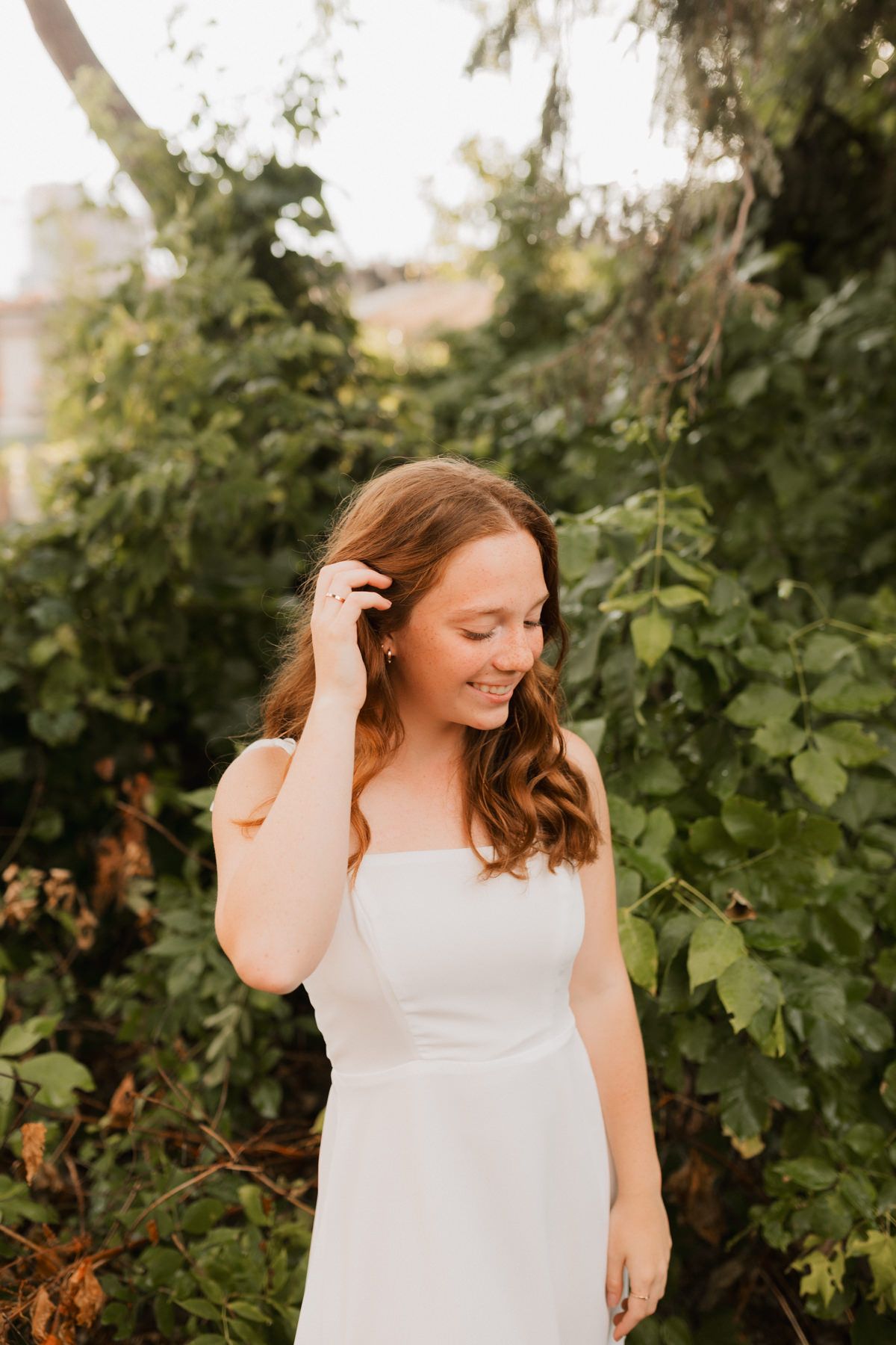 Young woman in white dress smiling outdoors surrounded by lush green foliage in golden hour light.