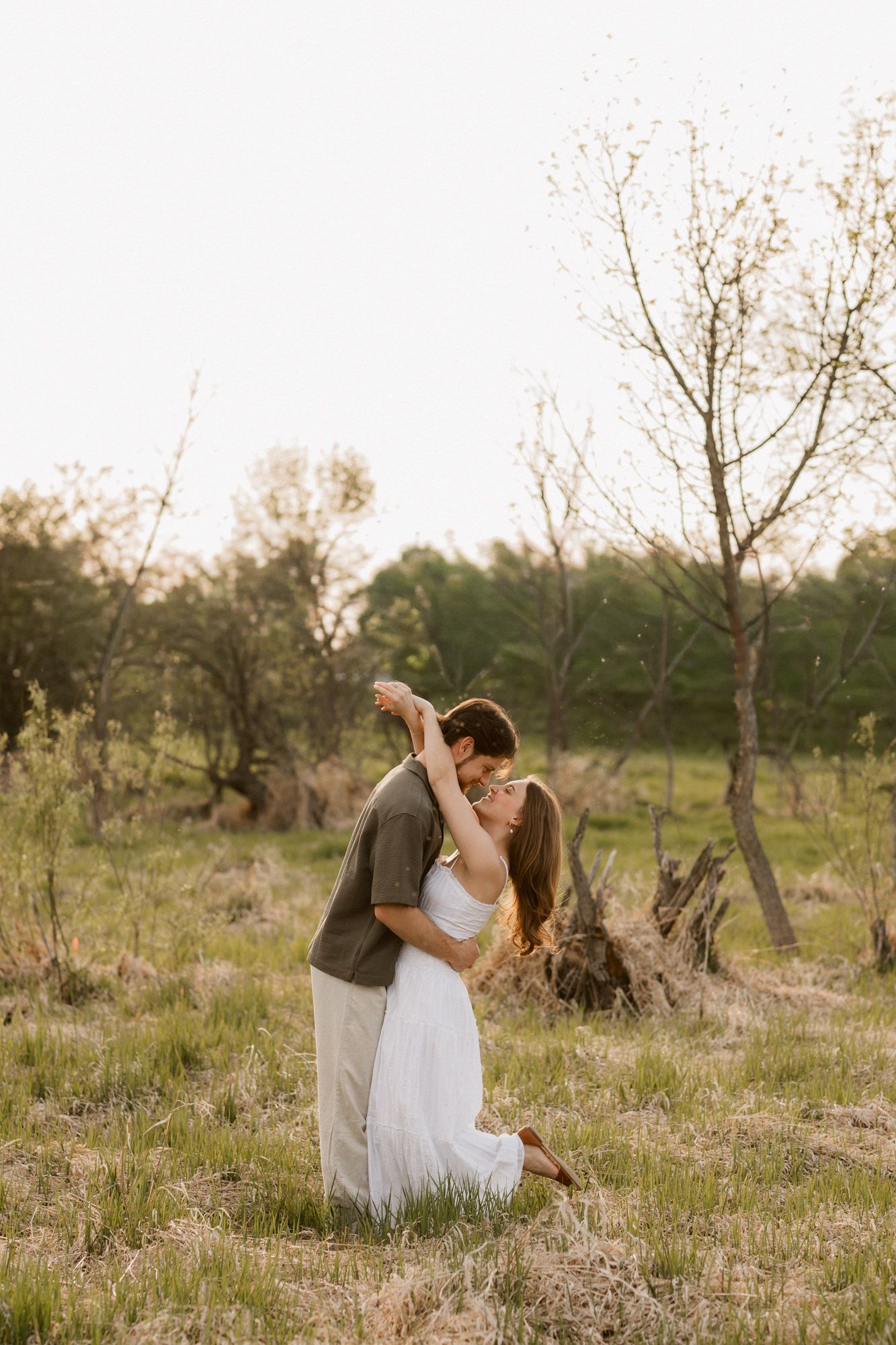 Couple sharing a romantic dance kiss in a sunlit meadow, woman in white dress, man in olive shirt, surrounded by trees.
