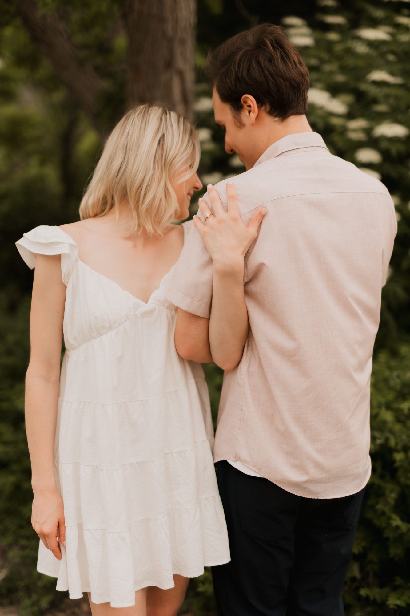 A couple shares a tender moment outdoors, woman in white dress touching man's shoulder, showcasing an engagement ring.