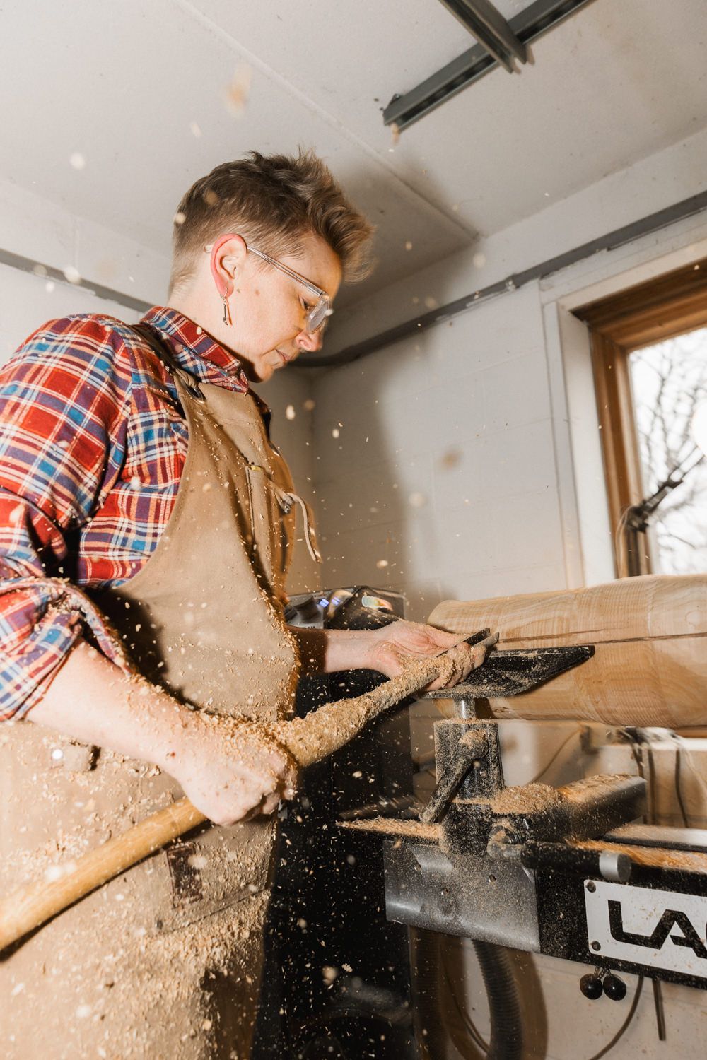 A woodworker in a plaid shirt and apron uses a power tool, sending sawdust flying in a workshop.