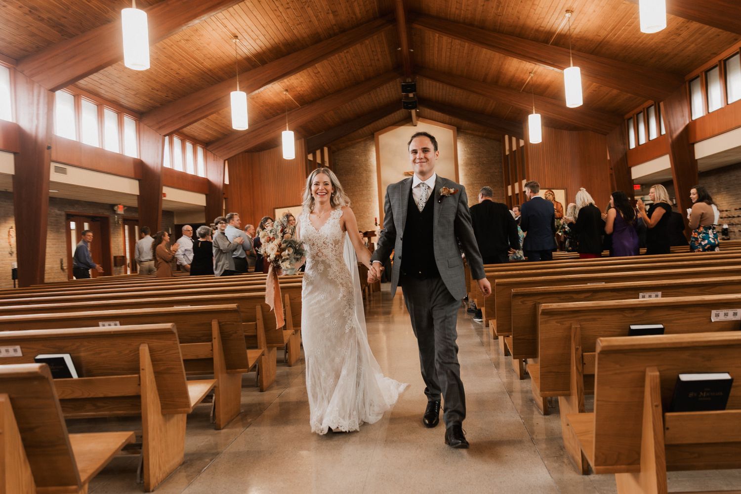 Joyful newlyweds walk down church aisle after wedding ceremony, bride in lace gown, groom in gray suit.