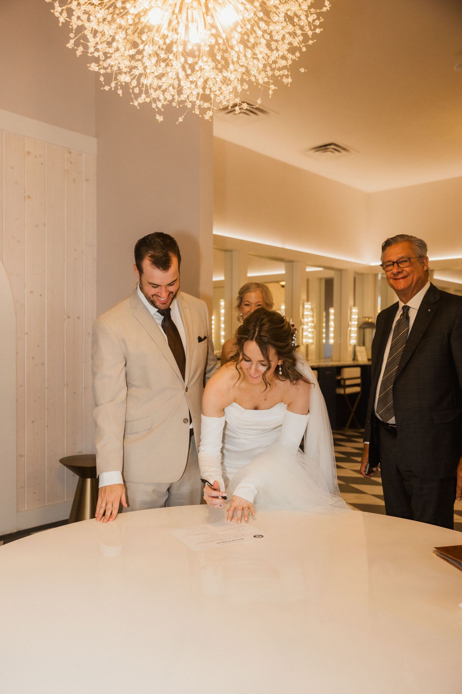 Bride signs the marriage license at a round table as the smiling groom and an officiant look on beneath a modern chandelier.