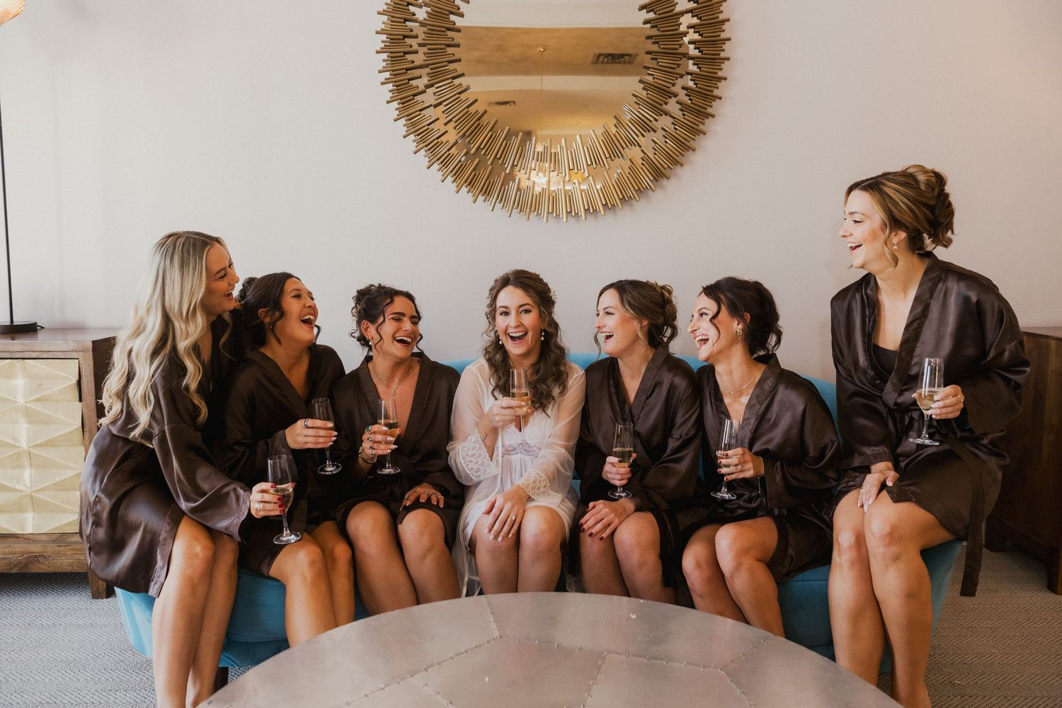 Seven smiling bridesmaids in matching satin robes laughing and holding champagne glasses while seated on a blue sofa before the wedding.