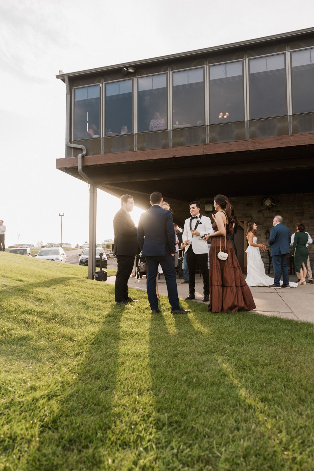Wedding guests gather outside a modern building on a grassy lawn during golden hour.