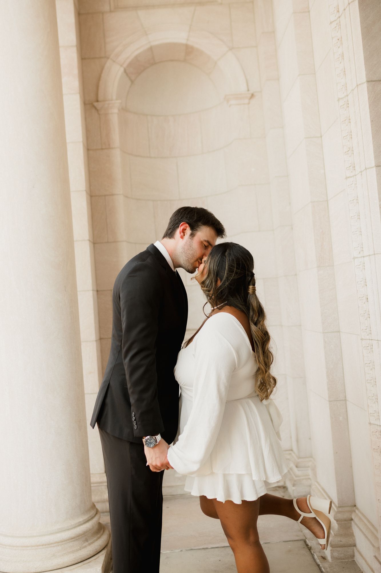 Couple sharing a romantic kiss between white marble columns, man in black suit and woman in white mini dress holding hands.