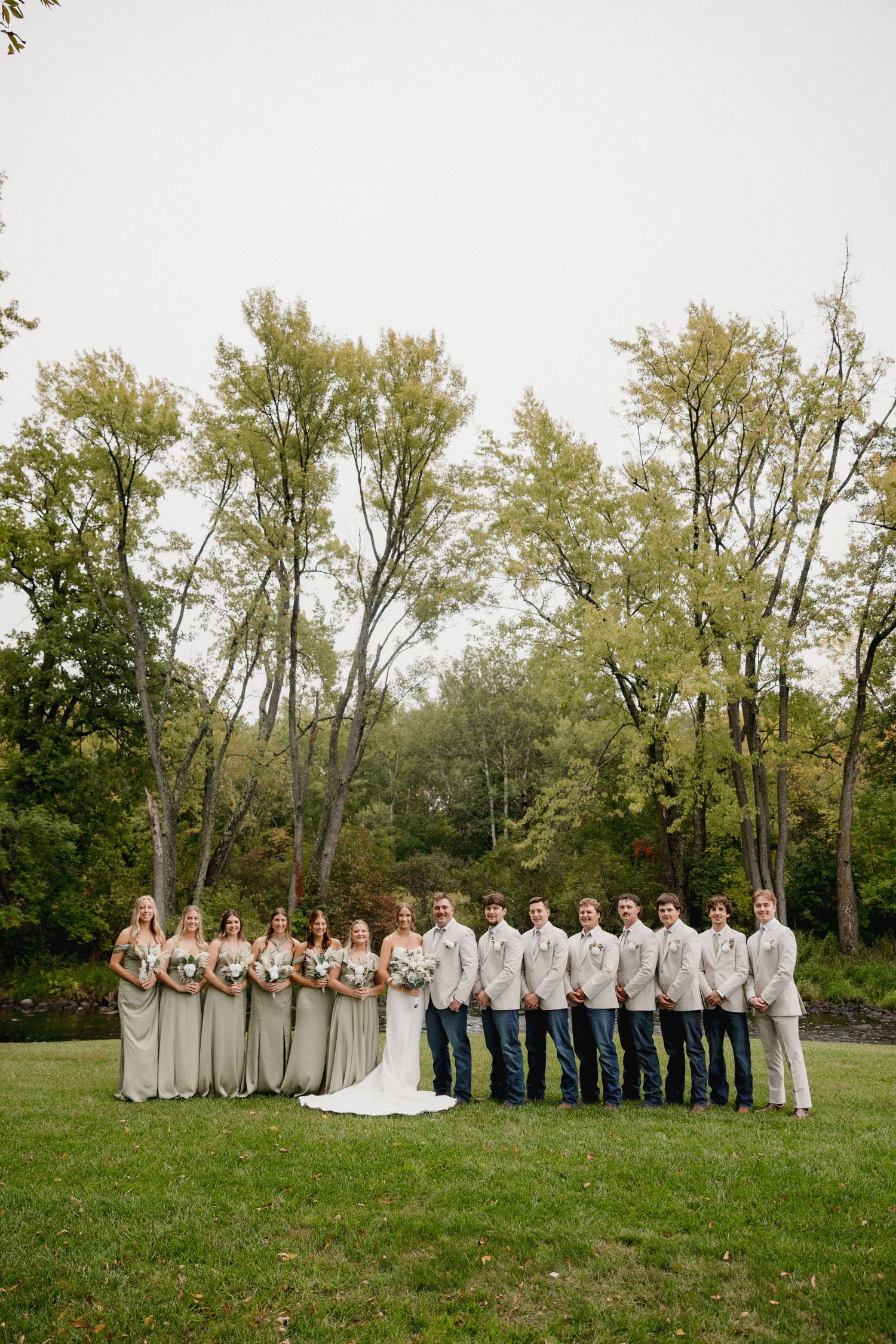 Wedding party poses outdoors on green lawn with trees, bridesmaids in sage gowns and groomsmen in jeans and tan suits.
