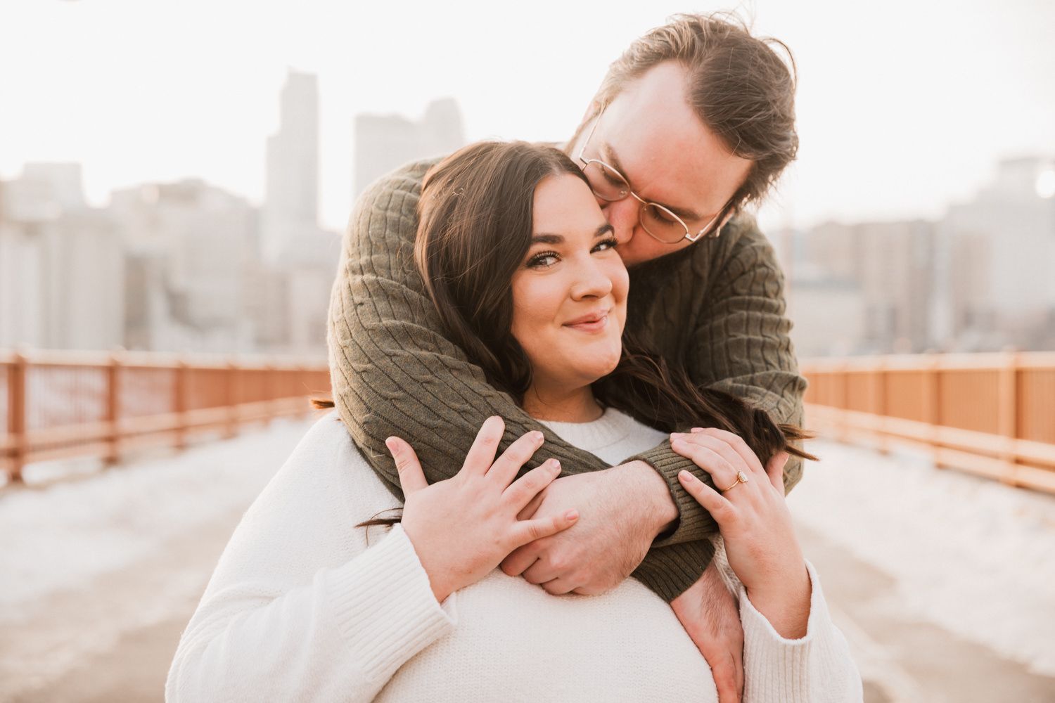 Romantic couple sharing tender moments on a bridge with urban skyline in soft golden light.