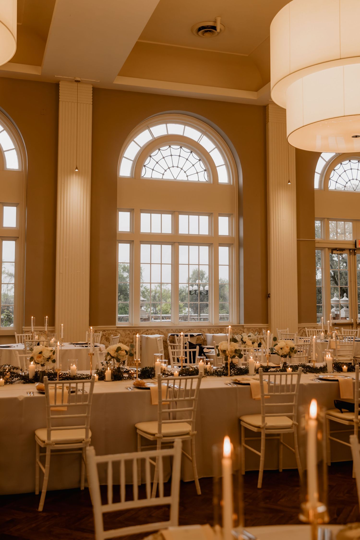 Elegant wedding reception hall with white chiavari chairs, long tables, candles, and large arched windows in a warm golden venue.