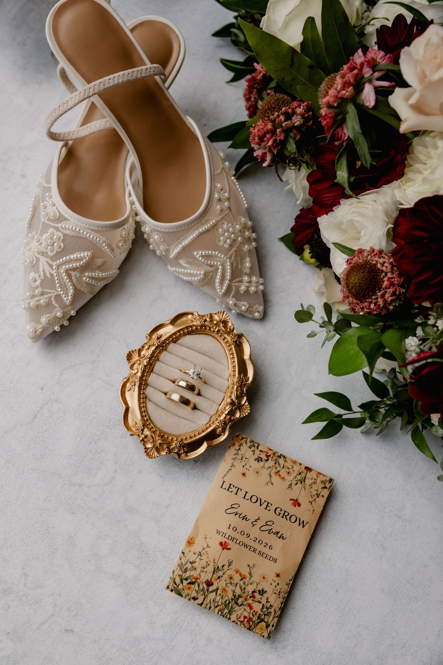 Elegant wedding flat shoes, gold ring holder, floral bouquet, and personalized seed packet favor on marble surface.