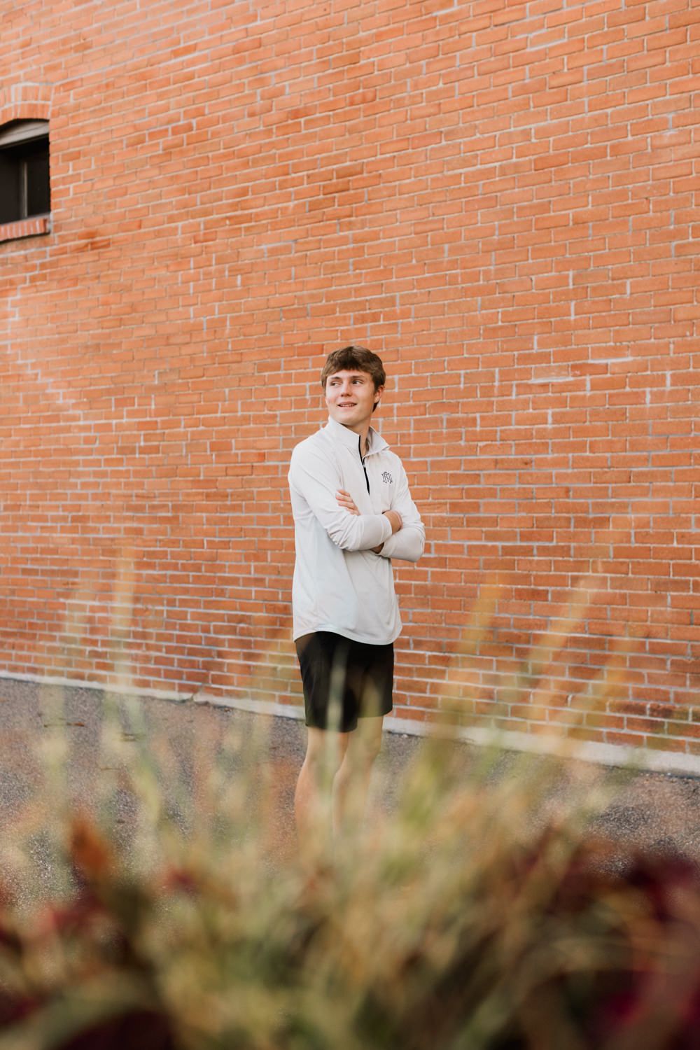 Someone in a white sweater poses against an orange brick wall on an autumn day with blurred foliage in foreground.