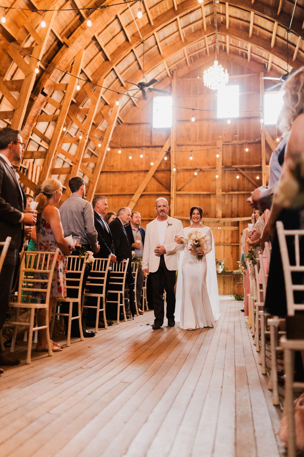 Guests line both sides of a wooden barn aisle during a wedding ceremony under vaulted ceilings.