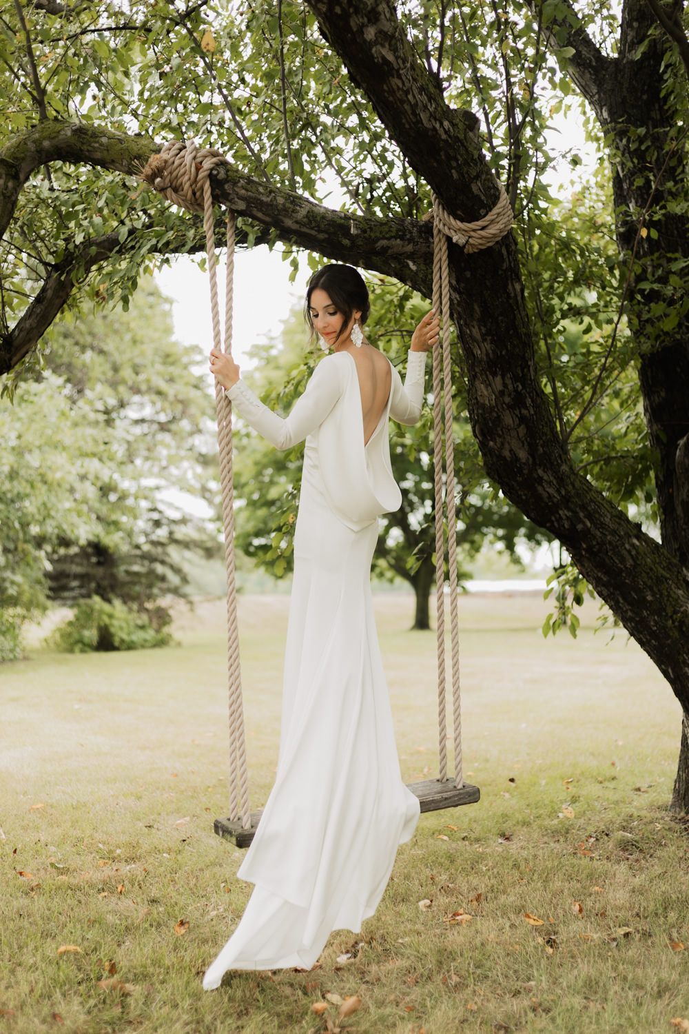 A person in an elegant white wedding dress stands gracefully on a rustic rope swing hanging from a large tree branch.