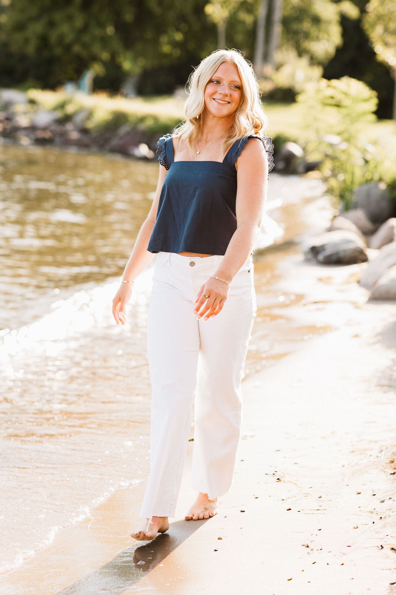 Woman in navy top and white pants poses gracefully by a waterfront on a sunny summer day.