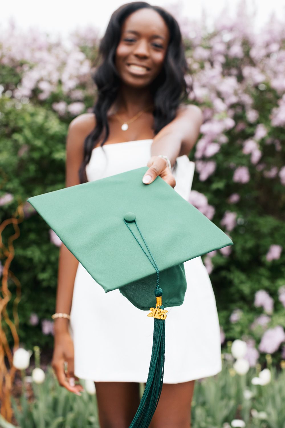 Graduate holds green mortarboard with tassel while wearing white dress against floral backdrop.
