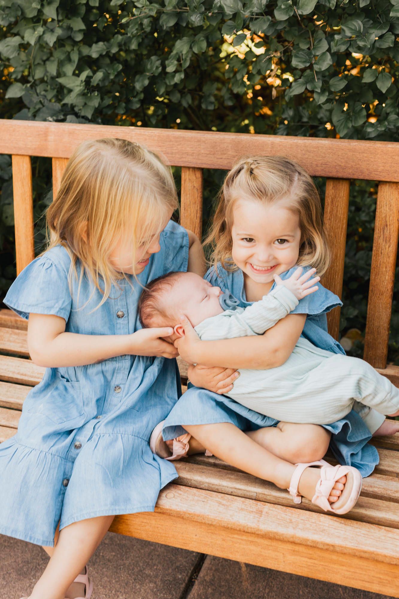 Children in matching blue dresses sitting together on a wooden bench with a small baby.