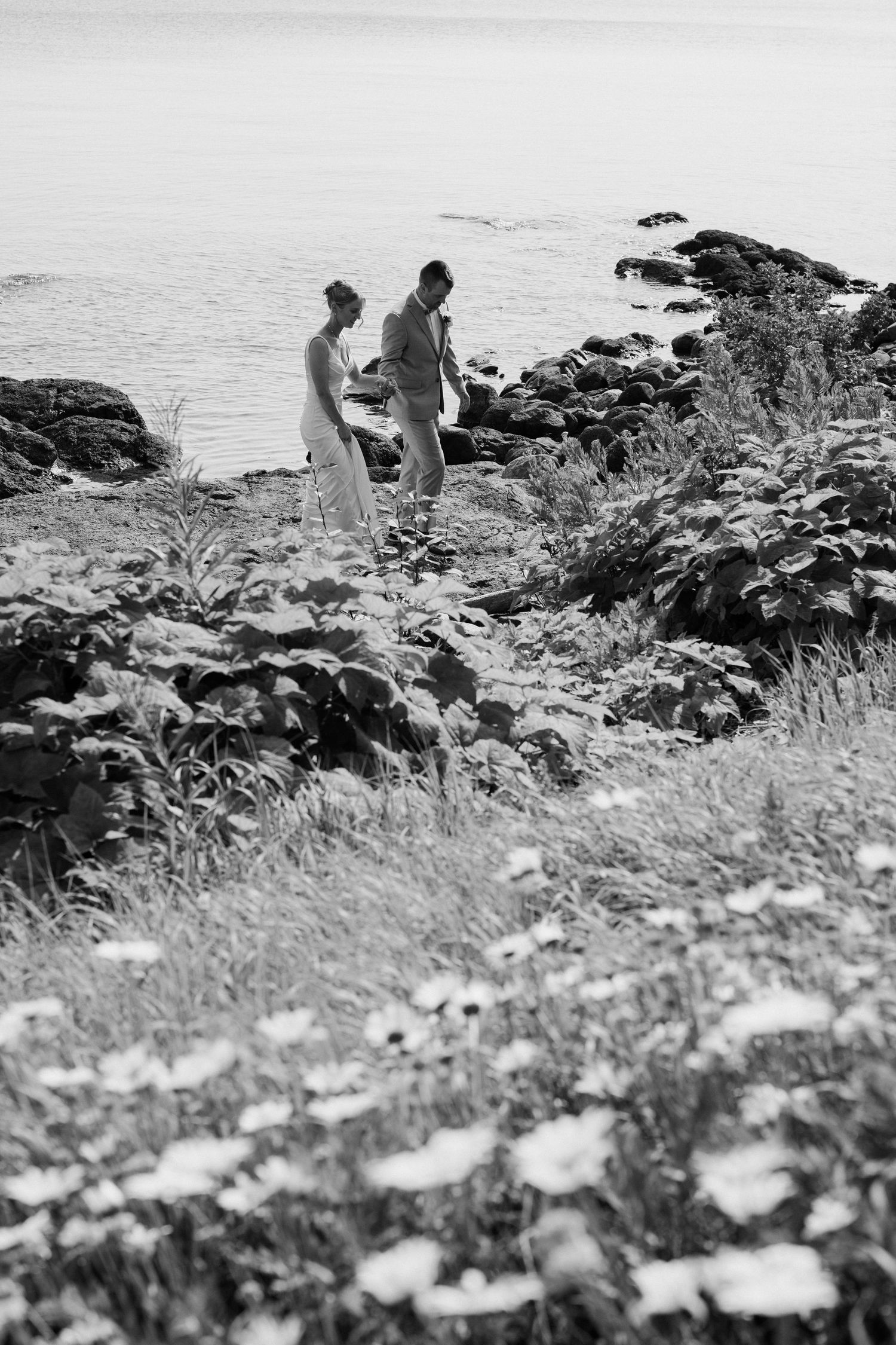 Black and white photo of a couple standing on a rocky coastal shoreline surrounded by wildflowers and lush greenery.