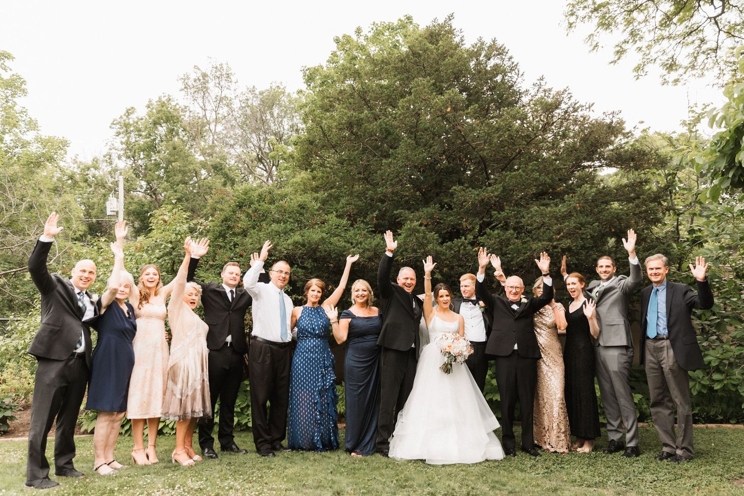 Wedding party celebrating outdoors, raising hands joyfully, bride in white gown surrounded by guests in formal attire.