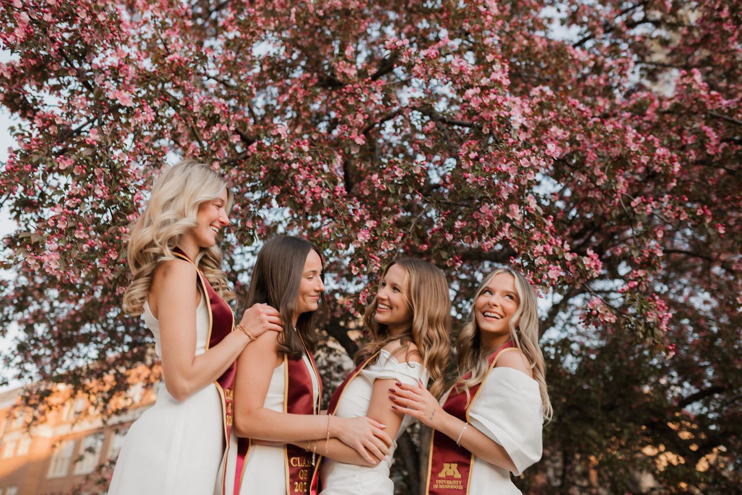 Three happy female graduates in white dresses with maroon stoles laugh under pink flowering tree.