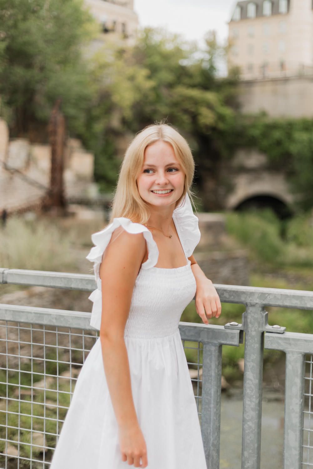 A person in a white summer dress poses on a metal railing outdoors against a blurred architectural background.