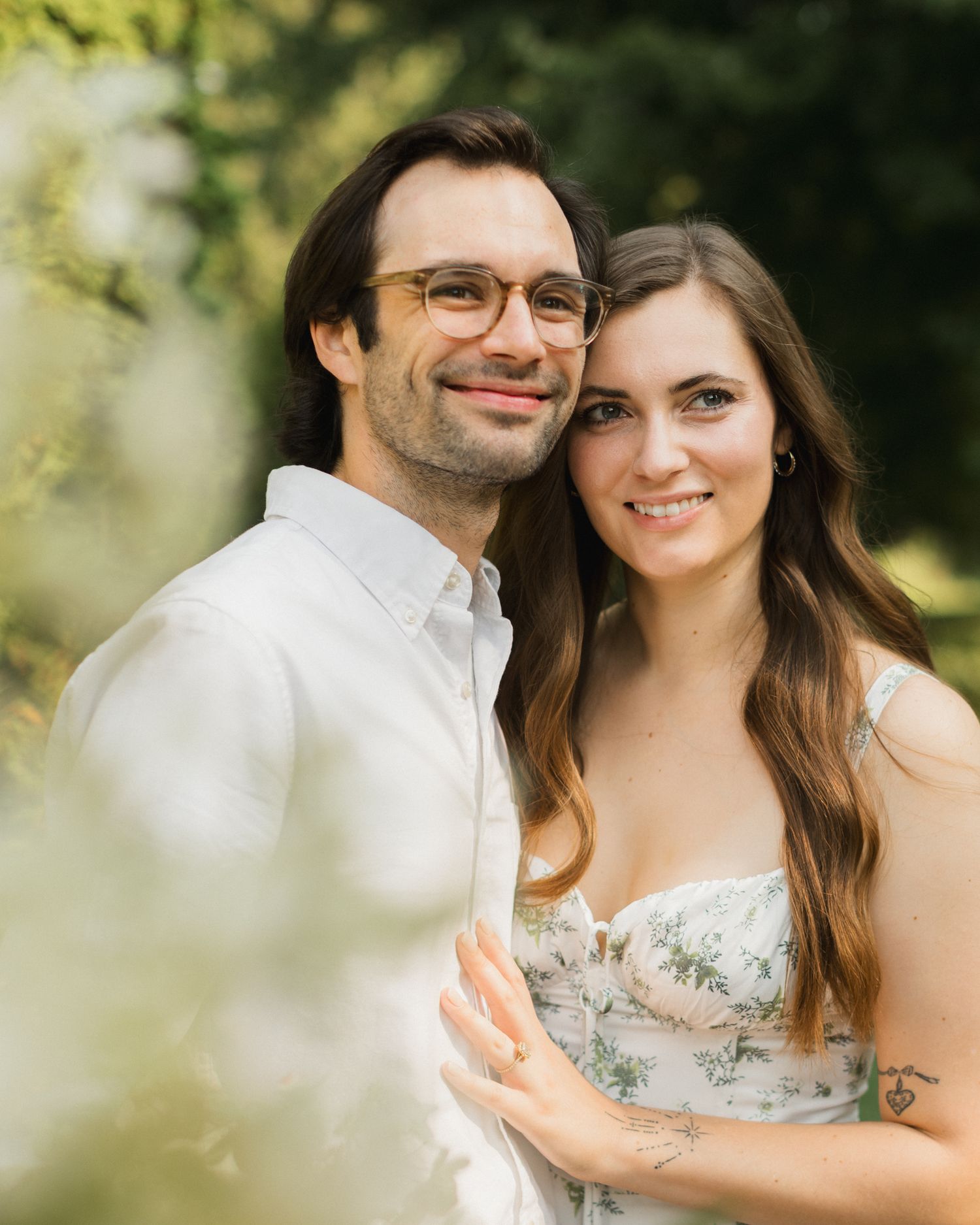 A smiling couple poses outdoors in soft natural light, man in white shirt with glasses, woman in floral dress.