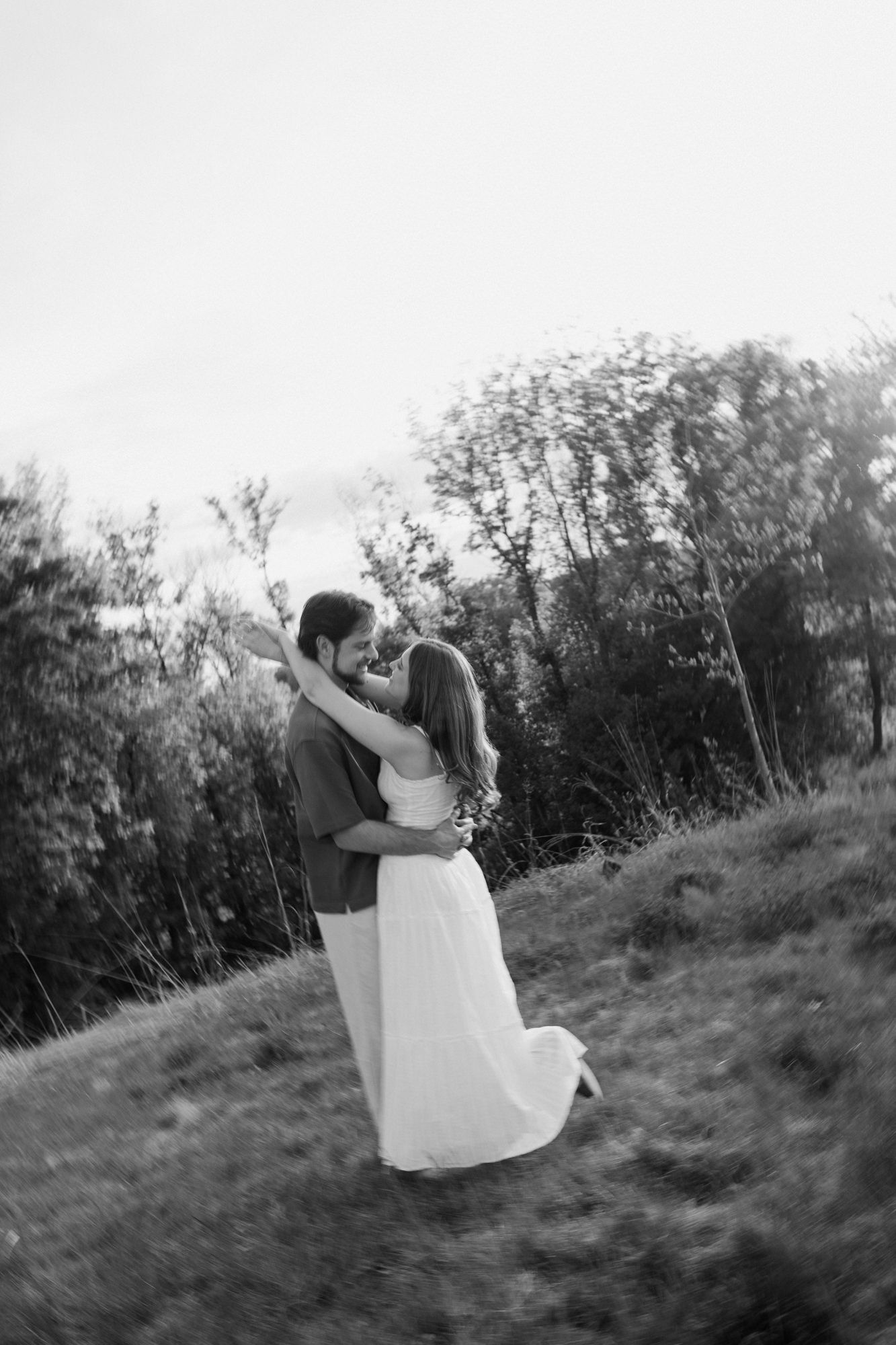 Black and white photo of a couple embracing and almost kissing on a grassy hillside surrounded by trees.