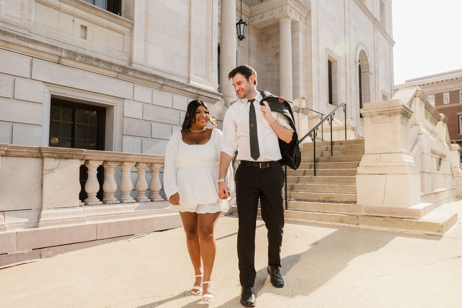 Interracial couple holding hands walking outside a grand building, woman in white dress, man in black suit.