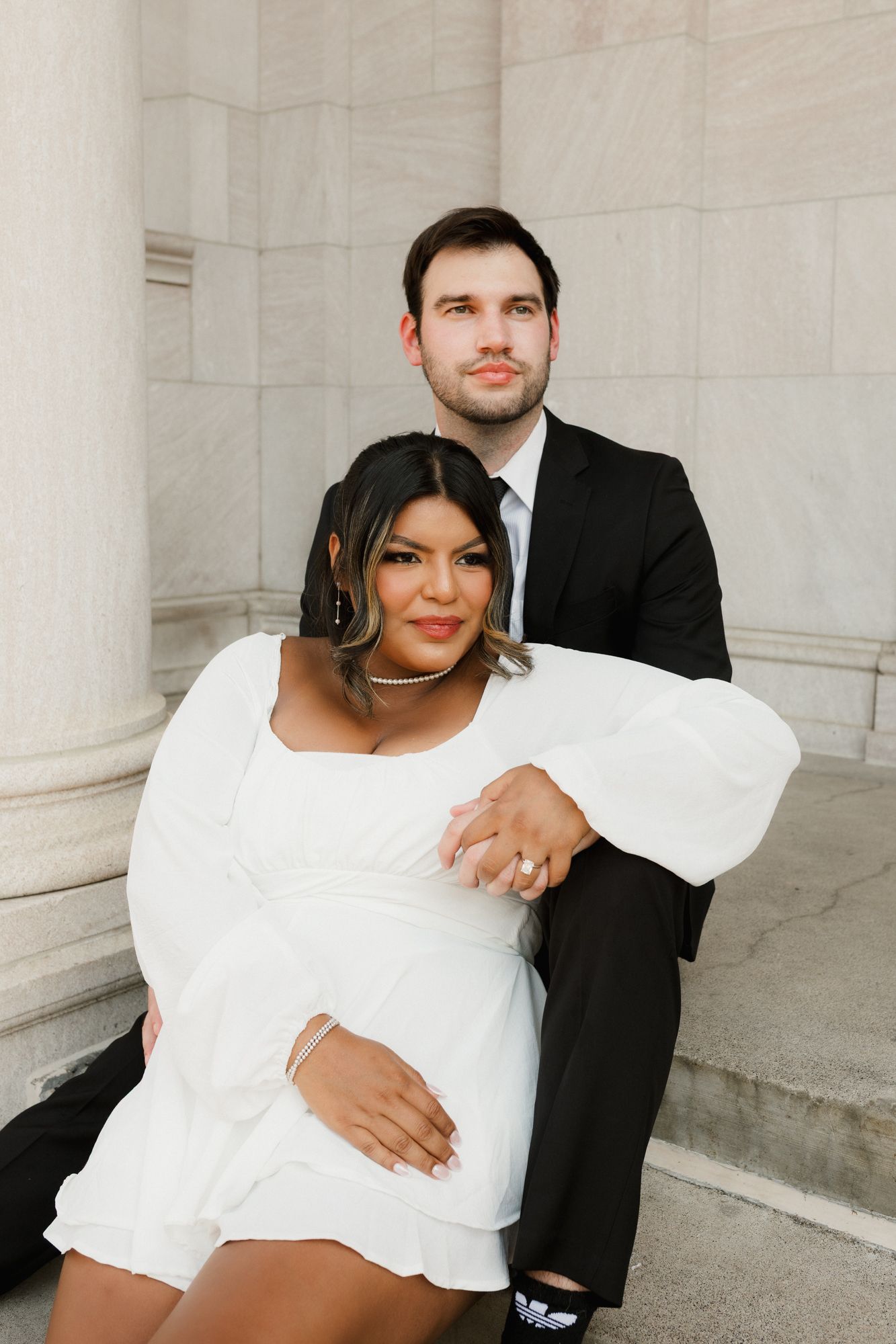 Couple posing on marble steps, woman in white dress cradling baby bump, man in black suit embracing her from behind.