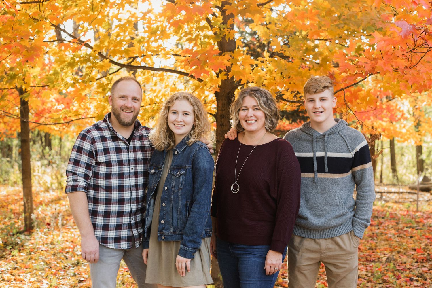 Family of four smiling outdoors among vibrant orange and yellow autumn foliage trees in a fall forest setting.