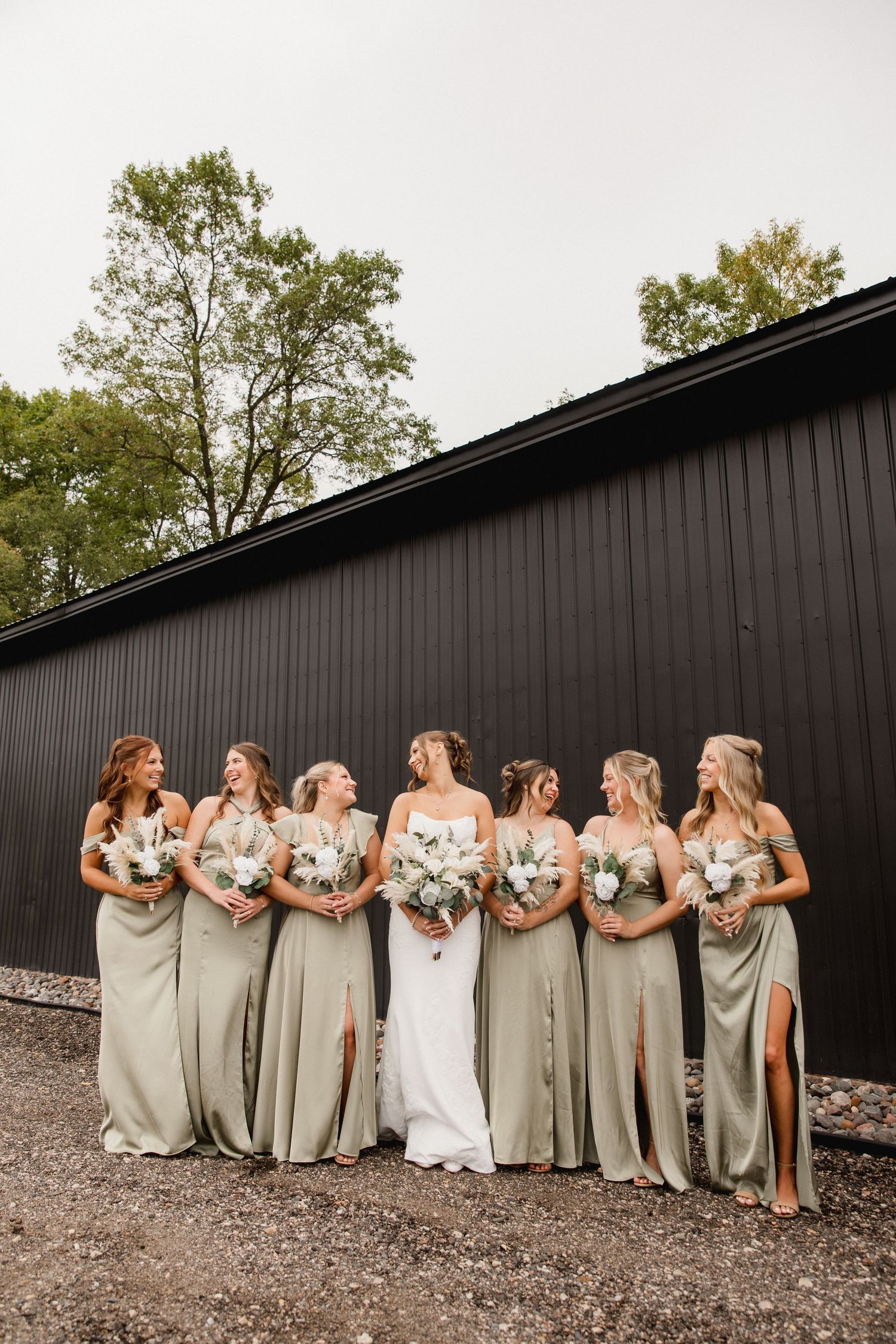 Bride in white gown with seven bridesmaids in sage green dresses holding white bouquets outside a black barn.