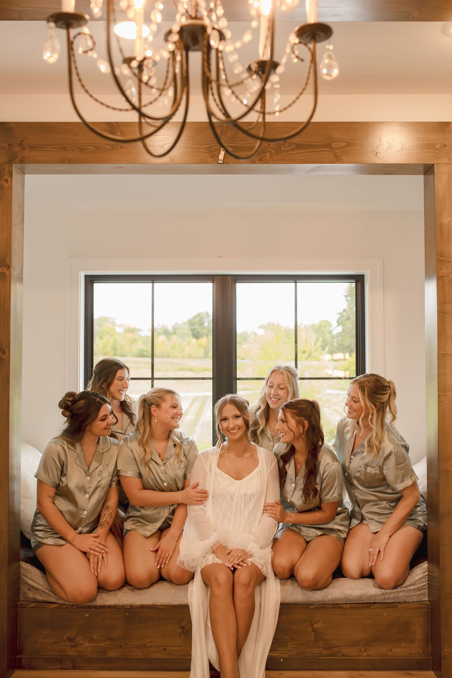 Bride in white robe surrounded by bridesmaids in sage satin sets, seated in a rustic window nook with chandelier.