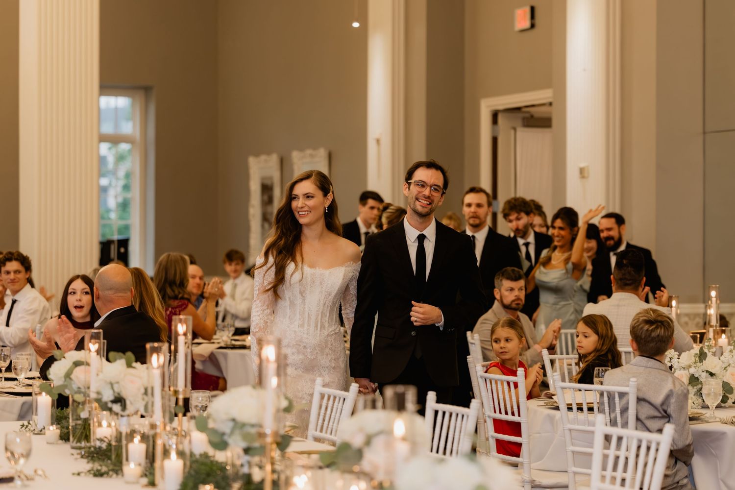Bride and groom make grand entrance at elegant wedding reception with candlelit tables and smiling guests.