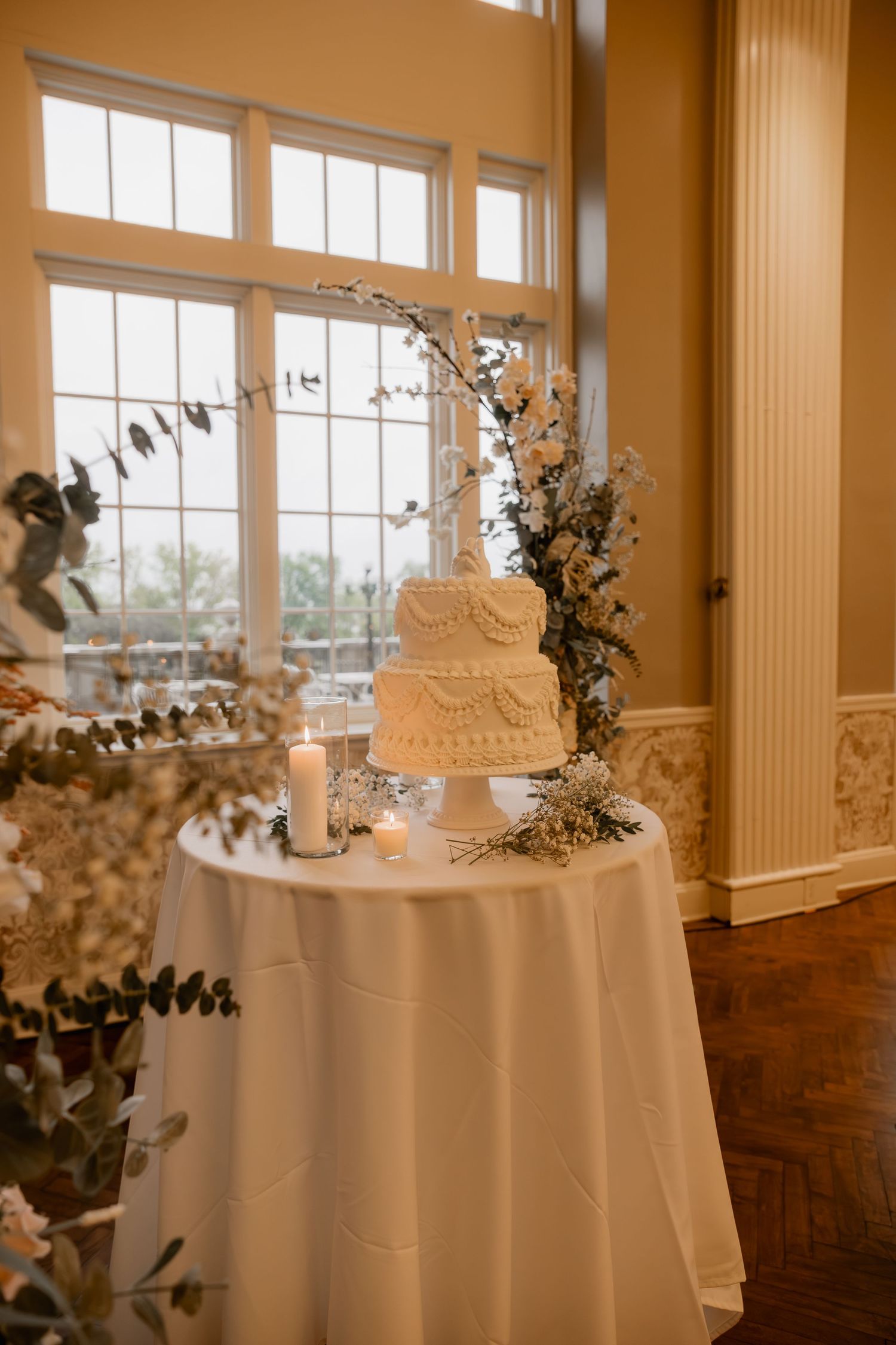Two-tier white wedding cake on draped table with floral arrangements and candles near large windows.