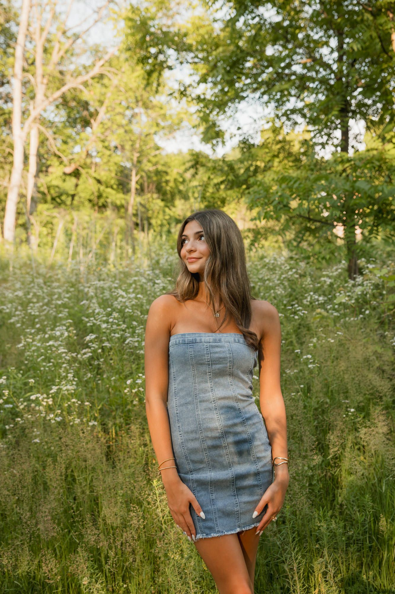 Young woman in denim tube dress standing in a lush green meadow with wildflowers and trees in the background.