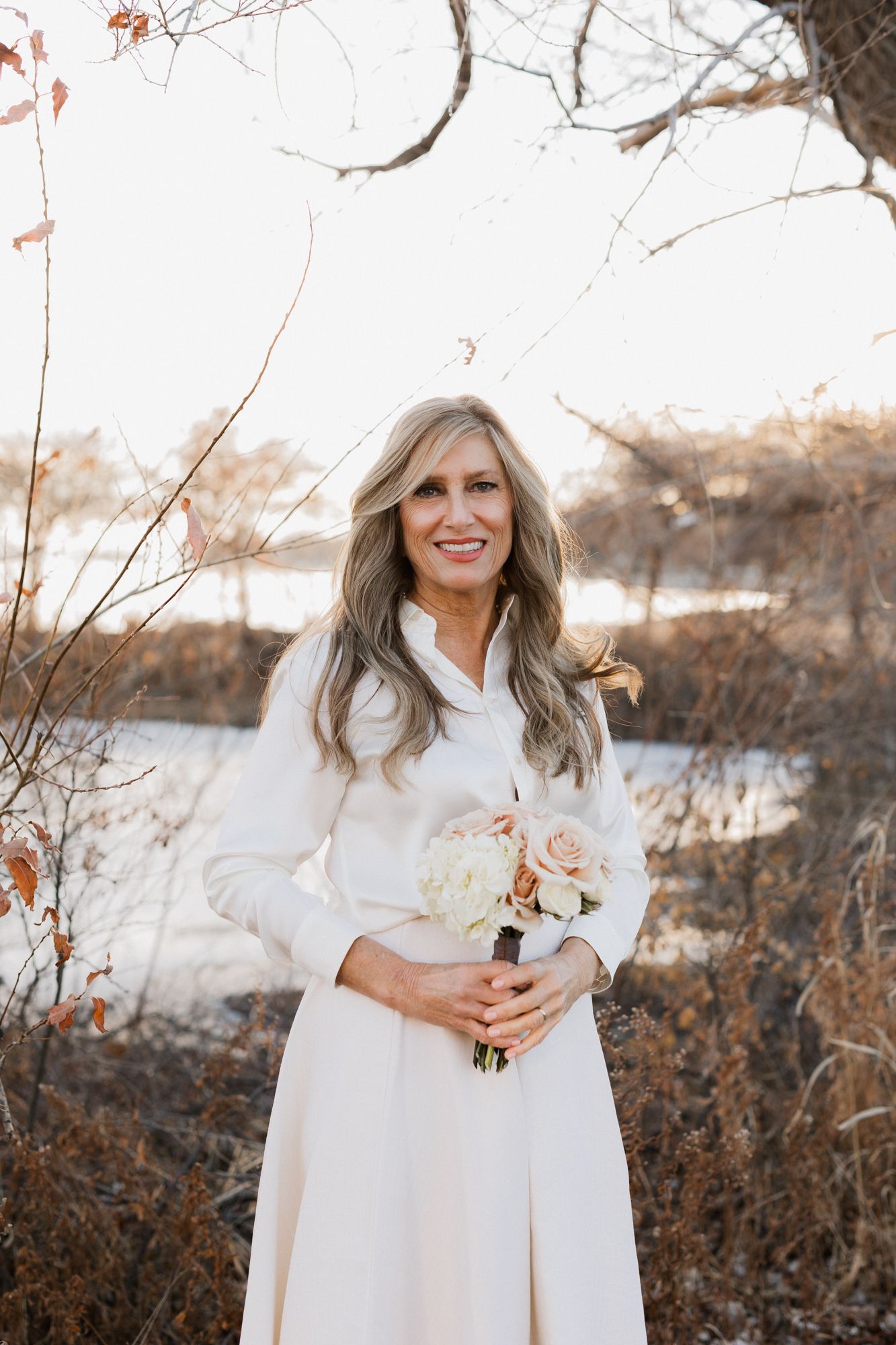 Smiling bride in white dress holding blush bouquet outdoors in winter setting with bare trees and snow.