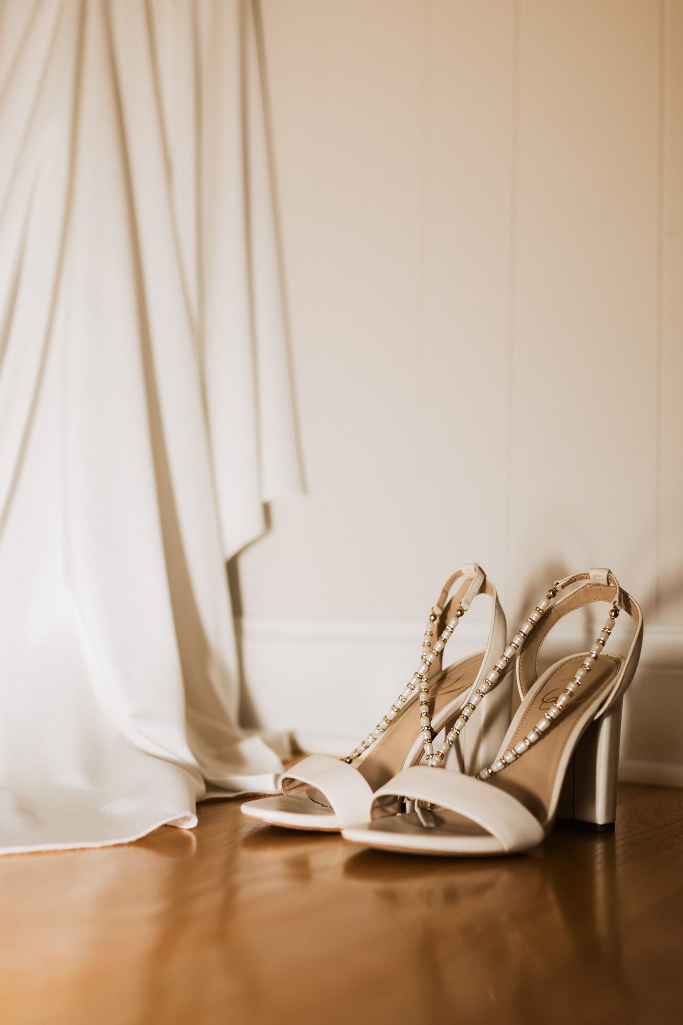Elegant white bridal heels with crystal embellishments on wooden floor beside flowing wedding dress.