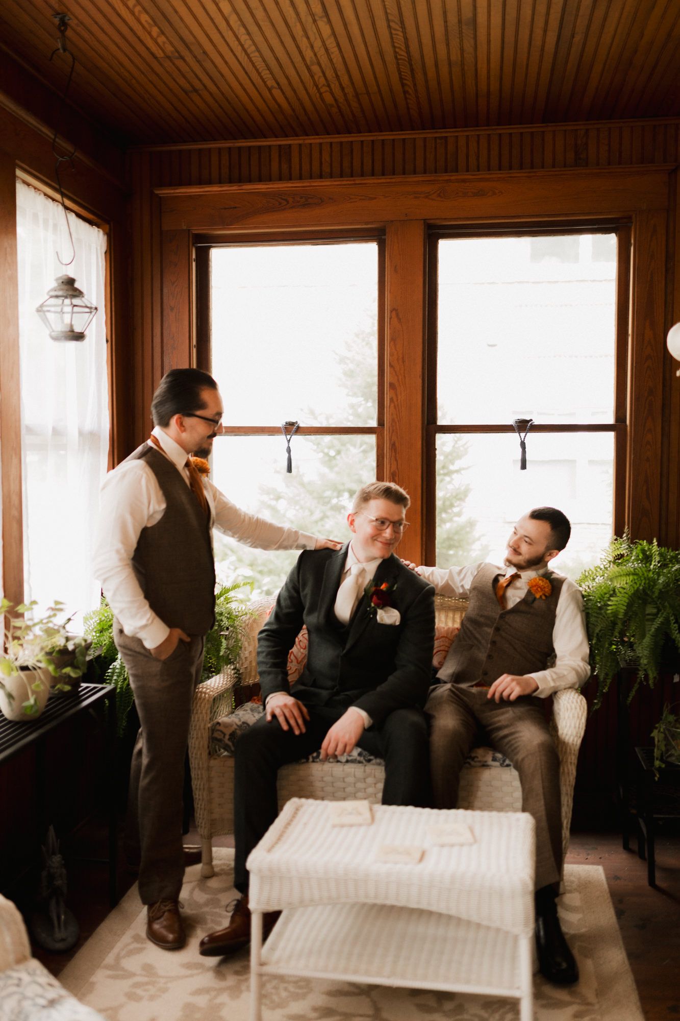 Three groomsmen in vests and boutonnieres relax in a wood-paneled sunroom before a wedding ceremony.