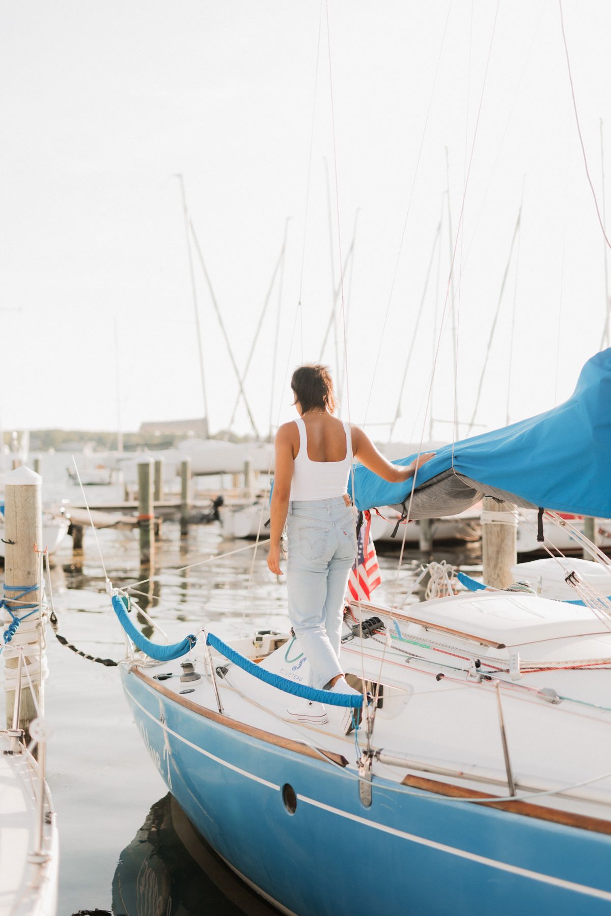 Woman in white top and jeans walking along dock beside blue sailboat at sunny marina.